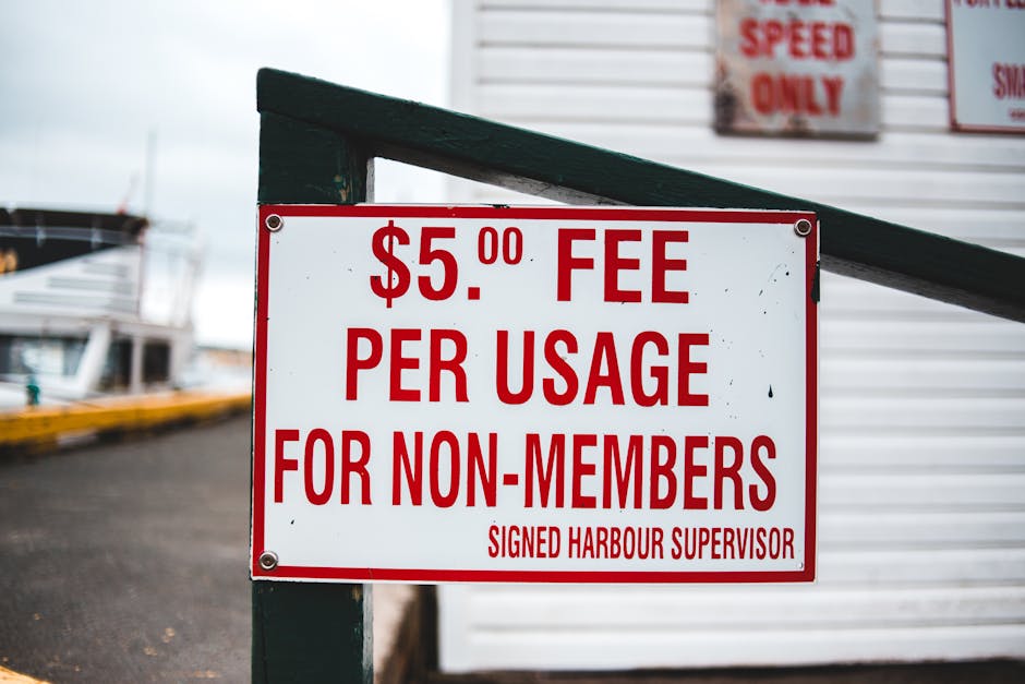 A white sign with red lettering displaying a fee of $5.00 per usage for non-members, attached to a dark green railing outside a marina or harbor area. In the background, blurred images of boats and a 