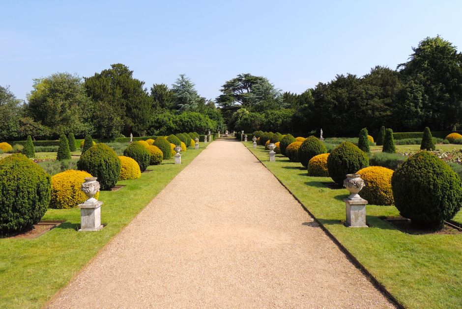 A wide gravel pathway runs through a formal garden with symmetrical, neatly trimmed round bushes and small classical urn-shaped stone planters lining both sides. Beyond the borders, there are well-mai